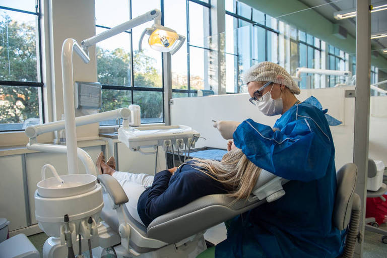 Vestida de macacão azul, aluna examina os dentes de paciente deitada em maca, sob luz de um refletor