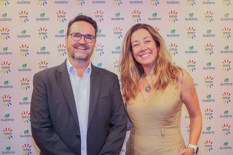 À frente de painel com logomarca do Instituto Coca-Cola Brasil e da Suzano homem e mulher posam para foto lado a lado. Ele usa blazer cinza e camisa azul-claro e ela usa vestido de cor beje. Ambos são pessoas brancas e sorriem