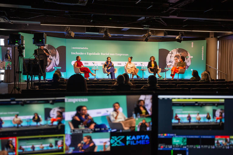 Foto ampla do palco com quatro convidadas e a mediadora; mais próximos da câmeras mas desfocados estão monitores de transmissão da equipe técnica