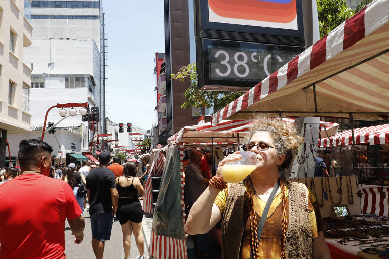 A imagem mostra uma mulher bebendo suco em uma feira ao ar livre. Ela está usando óculos de sol e um colete marrom. Ao fundo, há várias barracas com toldos listrados de vermelho e branco, e muitas pessoas caminhando pela rua. Um termômetro digital em um prédio ao fundo mostra a temperatura de 38°C.