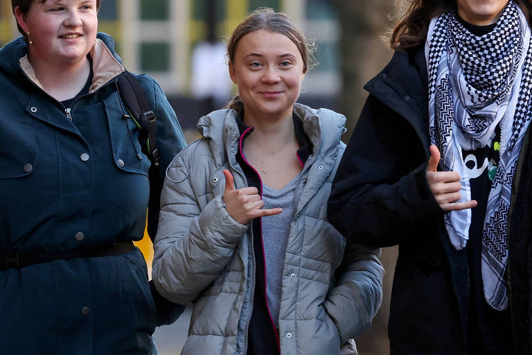 Greta faz um hangloose com uma das mãos enquanto caminha ao lado de duas pessoas