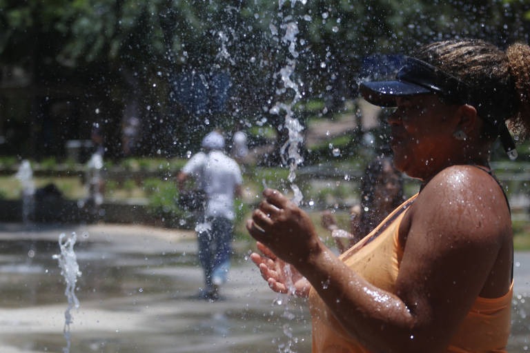 mulher se refresca com água que jorra de baixo para cima na imagem, há outras pessoas caminhando ao fundo e faz sol