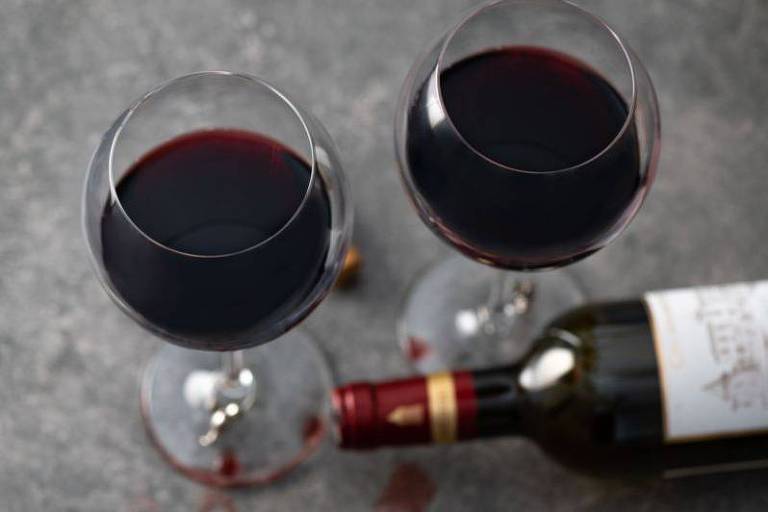 A imagem mostra duas taças de vinho tinto cheias, vistas de cima, colocadas sobre uma superfície cinza. Entre as taças, há uma garrafa de vinho tinto deitada, com a rolha ao lado. Há algumas gotas de vinho derramadas ao redor da garrafa.
