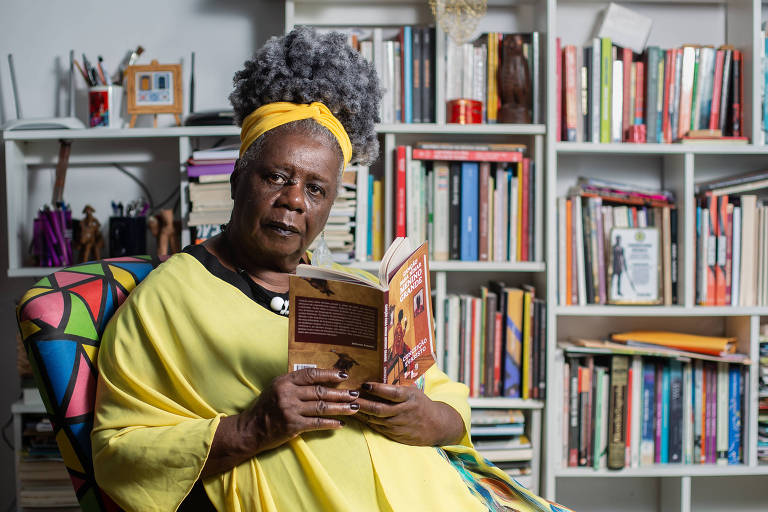 escritora negra de vestido amarelo e cabelo grisalho sentada com livro aberto na mão em frente a estante de livros