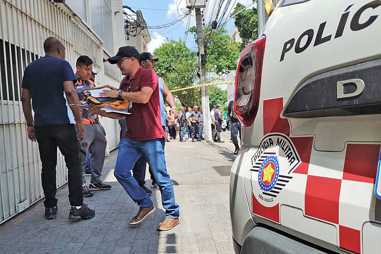 Homem com camisa vermelha e boné caminha com pacote nas mãos em direção a portão; em primeiro plano, escudo da Polícia Militar gravado na pintura de uma viatura