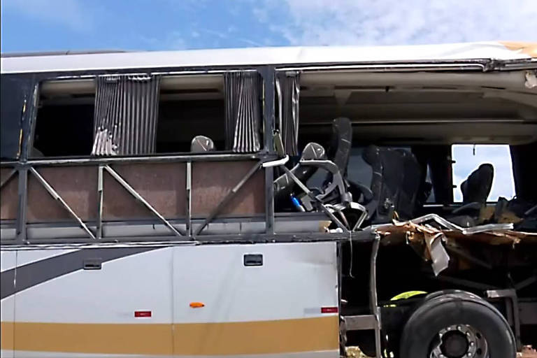 A imagem mostra um ônibus com danos significativos na lateral, onde a parte da frente está destruída e as janelas estão quebradas. As cortinas estão penduradas e alguns assentos são visíveis através da abertura. O céu está claro ao fundo