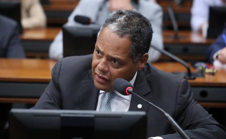 Foto mostra homem negro, de cabelo grisalho, falando ao microfone