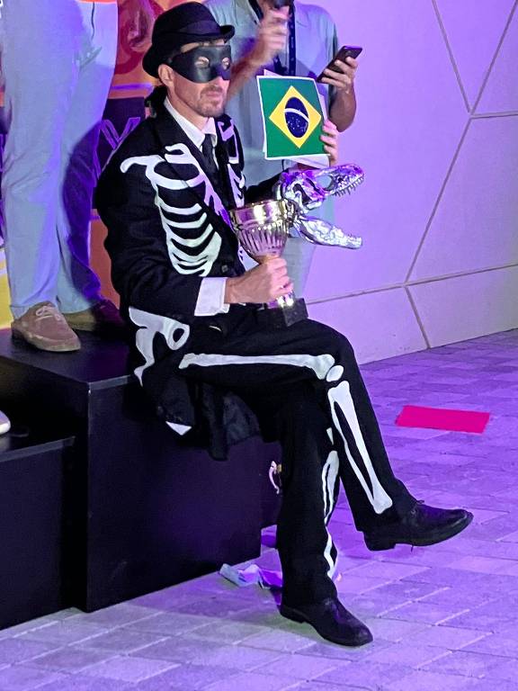 Homem com máscara segura troféu do Fóssil do Dia ao lado da bandeira do Brasil