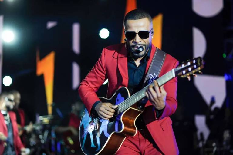 Em foto colorida, homem de terno vermelho toca violão em show