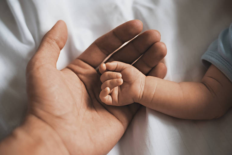 Palma de uma mão masculina voltada pra cima ampara uma maozinha de bebê