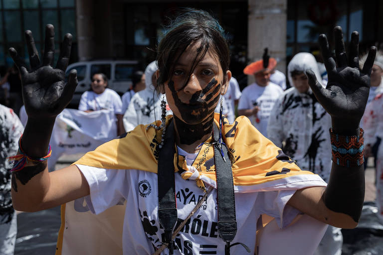 Indígena com camiseta branca, bandeiras amarelas e rosto pintado de preto abre os braços