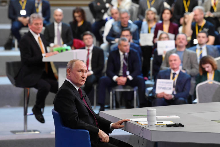 Observado pelo mediador e porta-voz Dmitri Peskov (na cadeira ao fundo) e com plateia de jornalistas com cartazes indicando seu meio de comunicação, Putin participa de entrevista anual em Moscou