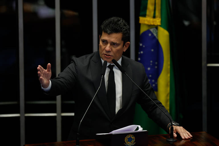 Moro veste terno e gravata, fala ao microfone e gesticula com uma das mãos, ao fundo bandeira do Brasil