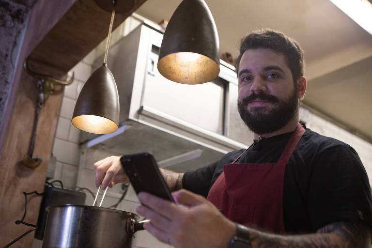 Homem branco, com cabelo e barba pretos usa camiseta preta, avental vermelho. ele olha para a foto e segura um celular em uma mão enquanto mexe com uma colher a comida em uma panela.