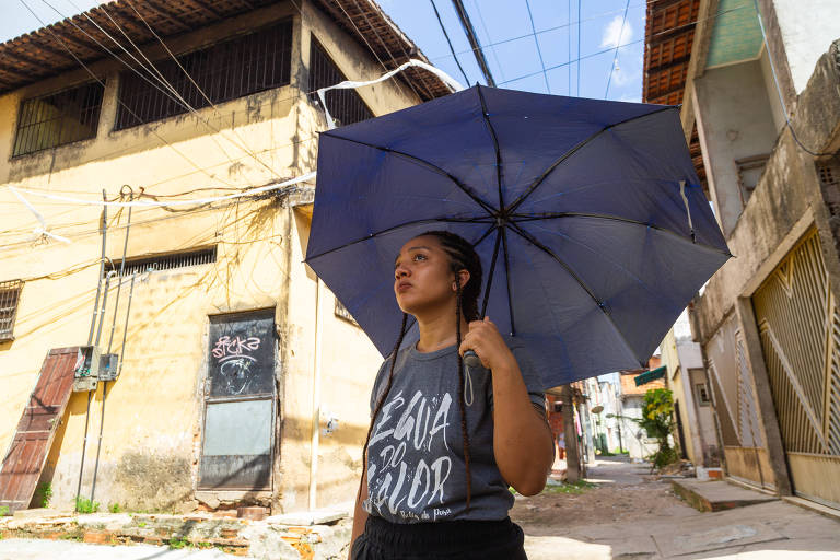 Mulher caminha com sombrinha em rua de casas de alvenaria sem reboco