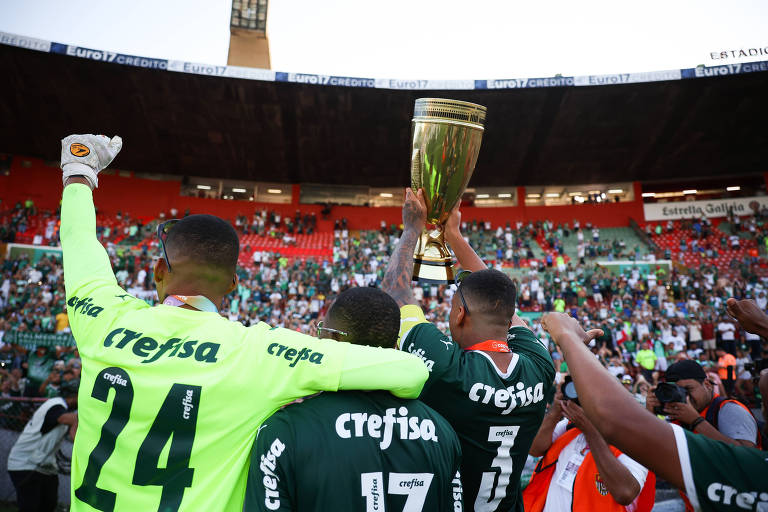 O goleiro, à esq., comemora abraçado com um companheiro de time, enquanto outro ergue a taça sobre a cabeça. Eles estão em frente à torcida