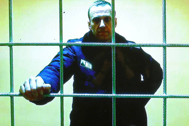Navalni é um homem branco e veste roupas de frio. Na imagem, ele está atrás de grades e segura uma das barras de ferro da cela.