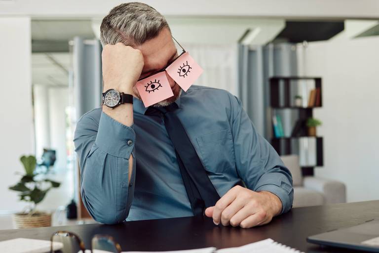 Fotografia colorida mostra uma homem branco sentado à mesa de um escritório, com post-its com desenhos de olhos abertos colados sobre as lentes dos óculos