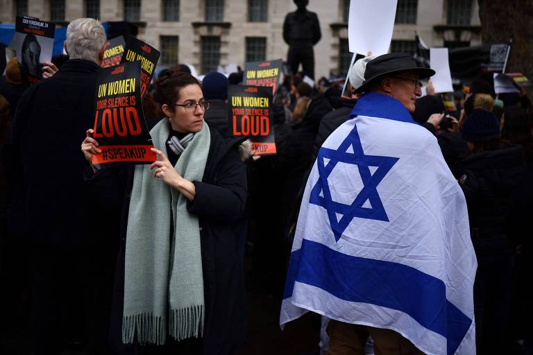 Mulheres exibem cartazes em que se lê 'ONU Mulheres, o seu silêncio é estrondoso' em protesto, enquanto um homem passa, enrolado na bandeira de Israel