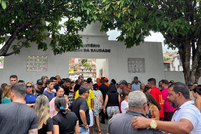 Dezenas de pessoas na porta do cemitério. Muro branco traz o nome "cemitério jardim da saudade"