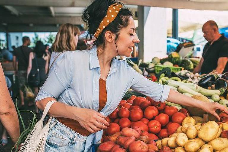 Mulher branca de cabelos pretos escolhendo frutas e legumes numa feira