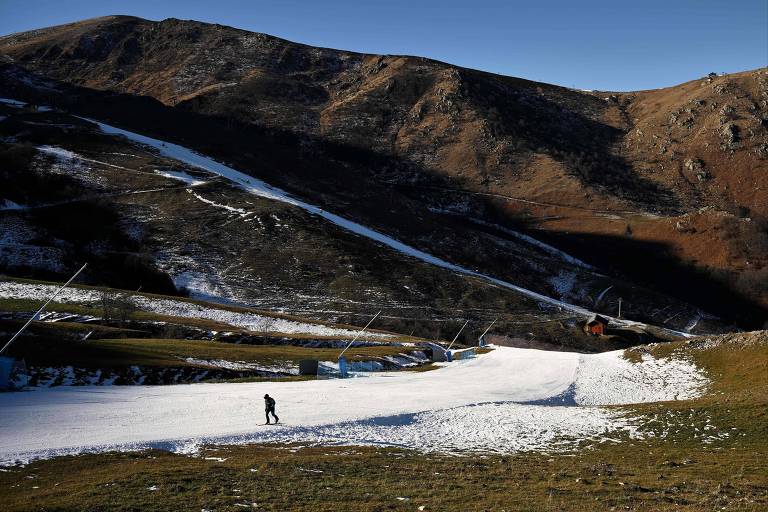 Pessoa caminha por pequena pista de neve cercada por montanhas completamente descongeladas