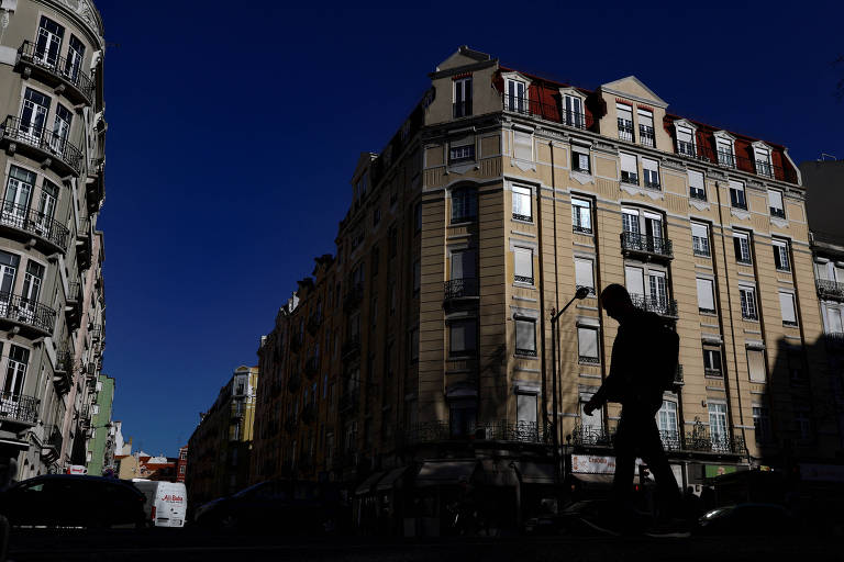 Pessoa caminha entre os prédios de Lisboa, capital de Portugal. Imagem mostra apenas a sombra de um homem. Ao fundo, um prédio maior, de cor bege. O céu está azul