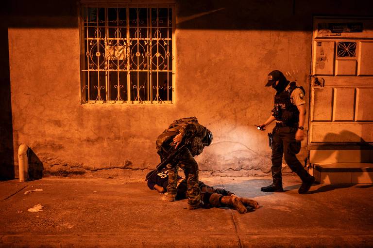homem deitado no chão de bruços é revistado por soldado armado, enquanto outro soldado observa, está de noite, há pouca iluminação