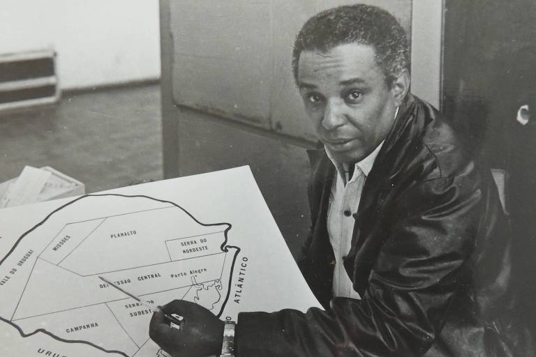 a foto mostra o cineasta odilon lopez apontando, com um lápis, para um mapa do Rio Grande do Sul que está em cima de uma mesa. ele é um homem negro retinto de cabelos crespos e curtos