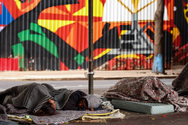 pessoas dormem com cobertores e sobre colchonetes na rua, há um muro colorido ao fundo 
