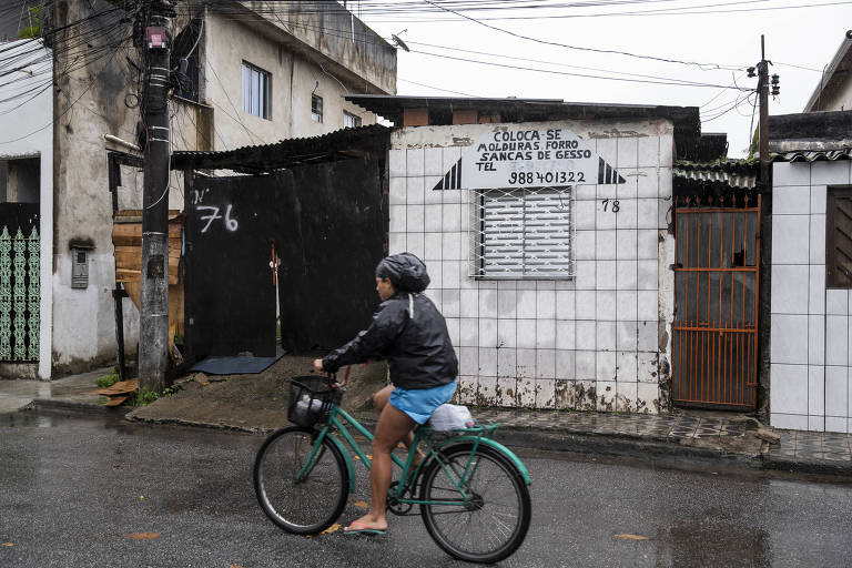 Homem com jaqueta e capuz em uma bicicleta azul sobre o asfalto molhado em frente a uma casa simples, com parede de azulejos brancos, janela persiana de alumínio e portão de grade laranja, onde há um cartaz anunciando molduras; ao lado há um portão de madeirite preta com o número 76 escrito com tinta