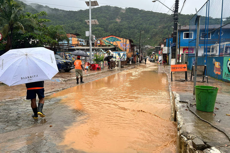 Pedrestras caminham por rua completamente alagada por água barrenta após temporal