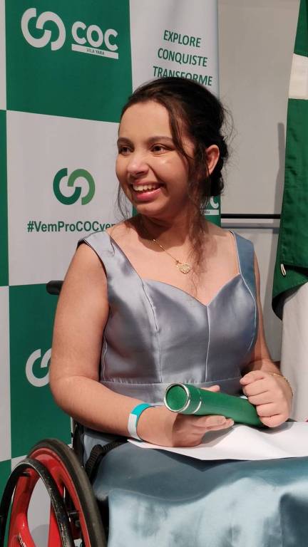 Uma jovem com um vestido verde prateado sorri enquanto segura um canudo de diploma. Ela está em uma cadeira de rodas em frente a um fundo verde e branco