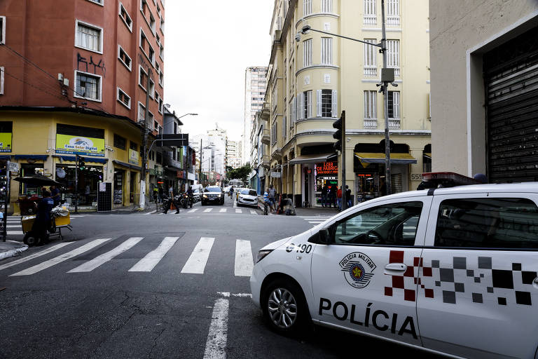 viatura do lado direito da foto está parada com a frente virada para a rua, ao fundo há pessoas circulando. a imagem é margeada por prédios dos dois lados