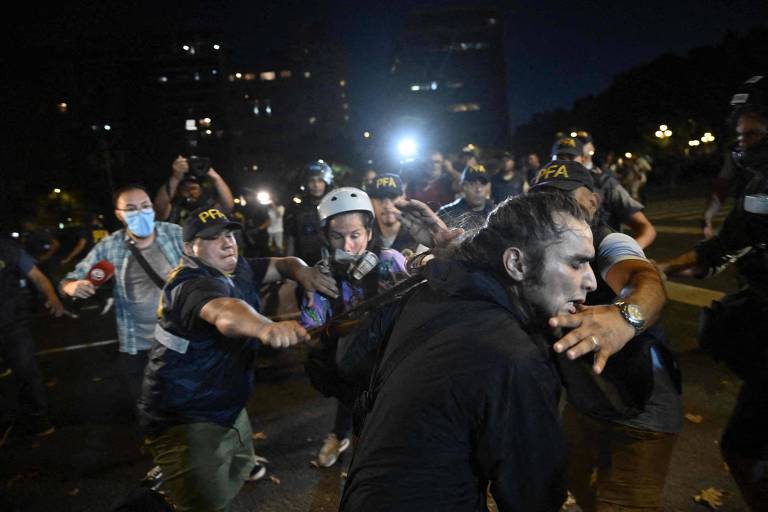 Foto noturna mostra homem de perfil, cabelo preto e rosto maquiado de branco, sendo detido por um policial, e manifestantes e policiais ao fundo