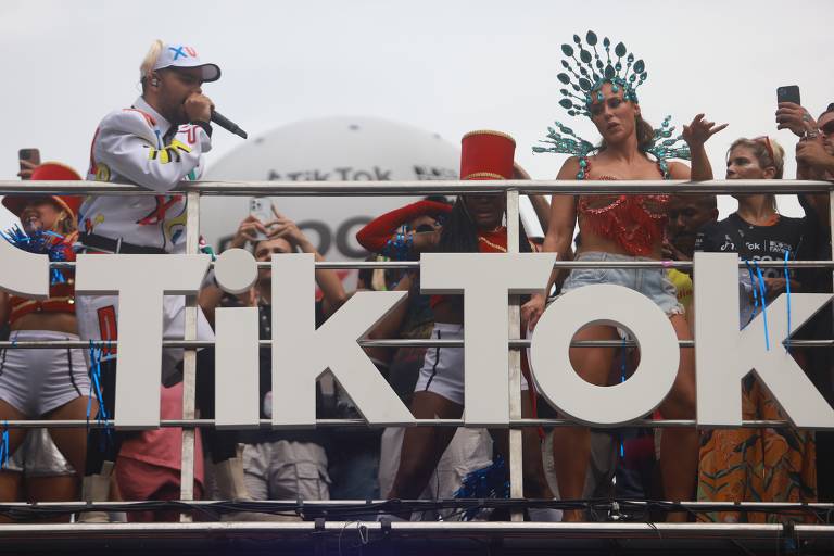 em cima de trio elétrico, pedro sampaio fala ao microfone, usa boné, e à esquerda, a atriz paolla oliveira samba, está fantasiada, há outras pessoas no trio; grande texto à frente da grade do trio diz "TIKTOK"