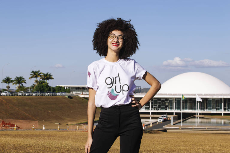 menina parda, de cabelos crespos, posa de camiseta branca e calça preta em frente a um gramado do congresso nacional, céu azul no fundo