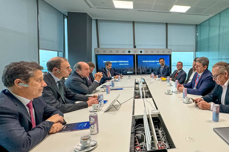 Homens de ternos sentados em mesa de reunião, com telas de apresentação ao fundo