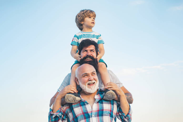 Fotografia de um senhor branco, calvo, olhando para o alto. Atrás dele, um homem branco de cabelos e barbas escuras olha na mesma direção. Sobre o ombro desse homem, um menino de aproximadamente cinco anos, também branco, com cabelos lisos e curtos. Os pés do garoto estão sobre os ombros do senhor mais velho.