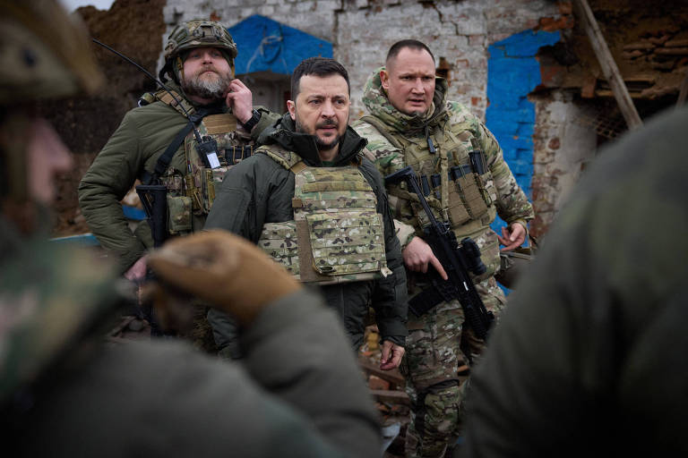 Foto mostra homem com barba e colete à prova de balas, o presidente da Ucrânia Volodimir Zelenski, cercado por soldados em uniformes verdes
