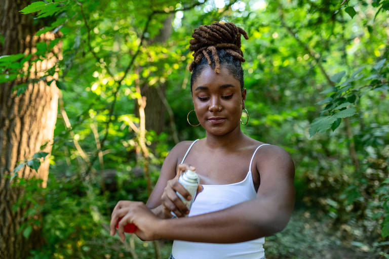 Uma mulher negra passa repelente no braço; no fundo da foto há muitas plantas