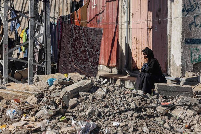 Mulher sentada, vestindo vestimenta islâmica preta, observa escombros 