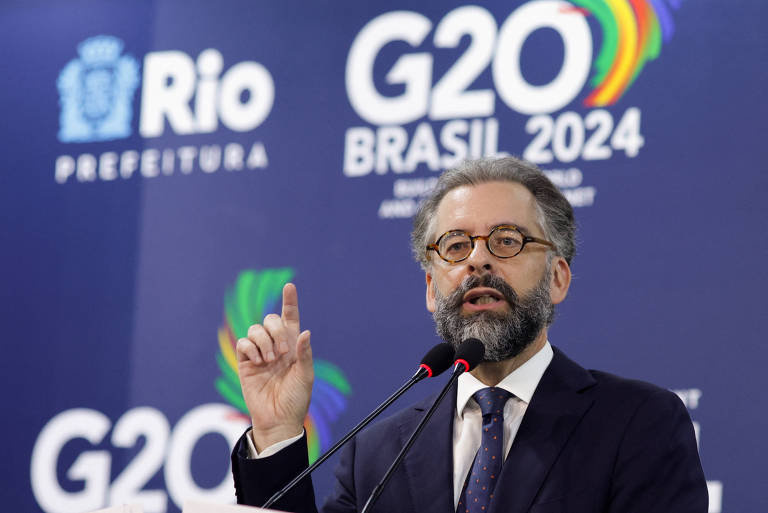 m homem de barba e óculos está falando em um microfone durante um evento. Ele está usando um terno escuro e uma gravata. No fundo, há um banner azul com os textos 'Rio Prefeitura', 'G20 Brasil 2024' e 'G20'.