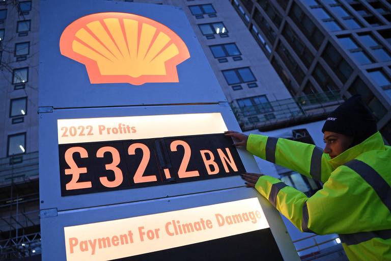Pessoa cola cartaz dizendo '32,2 bilhões de libras esterlinas' em um painel idêntico aos usados em postos de gasolina para indicar os preços dos combustíveis
