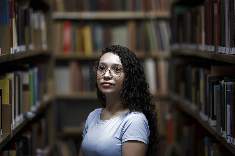 Jovem de óculos e cabelos encaracolados usa blusa azul e posa em meio a estante de livros