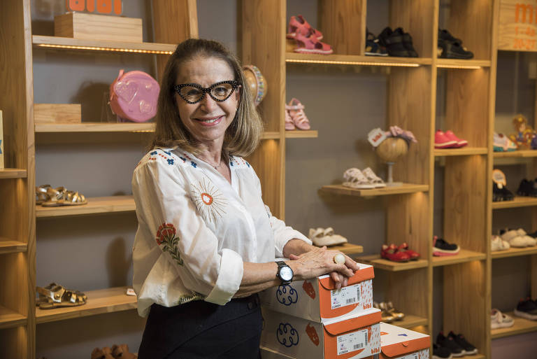 Sorrindo, uma mulher está de pé, dentro de uma loja de sapatos infantis, apoiada em uma pilha de caixas de sapatos. Ela usa um óculos com armação preta e veste uma camisa branca com alguns bordados