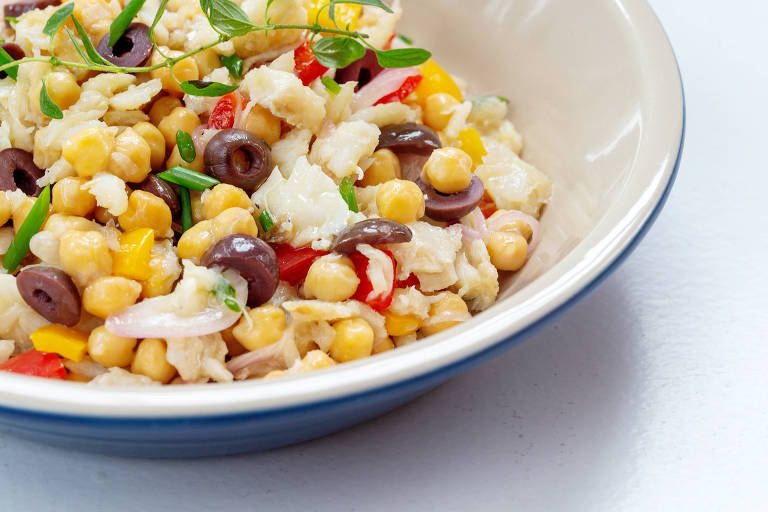 Prato de cerâmica com laterais brancos com salada bacalhau com grão-de-bico, com receita vista de cima; prato está em cima de superfície branca  


