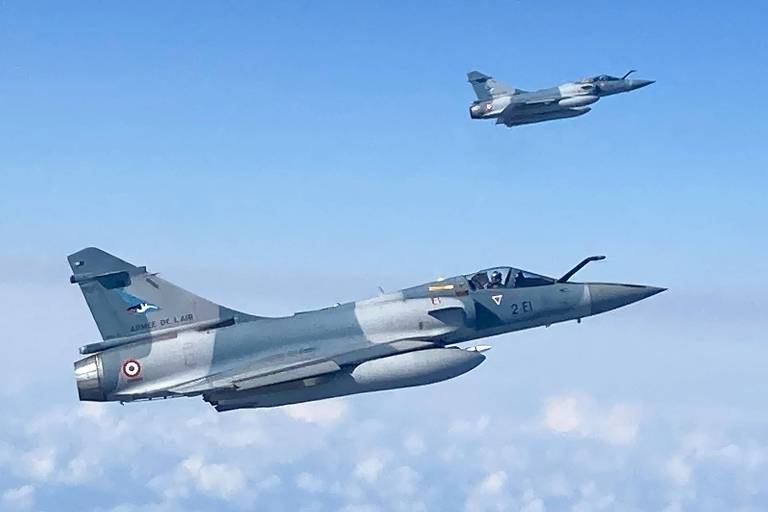 Dois aviões de caça cinza-azulados voam em um céu azul. Eles são Mirage-2000 franceses e patrulham a Estônia, no mar Báltico