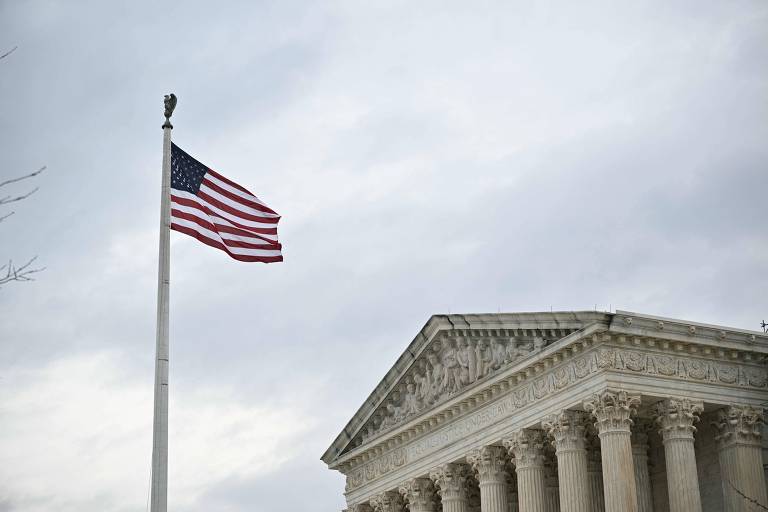 Foto mostra bandeira dos Estados Unidos flamulando ao vento sob um céu cinza. No fundo, é possível ver a parte de cima da sede da Suprema Corte americana, um prédio no estilo clássico com triângulo e colunas.