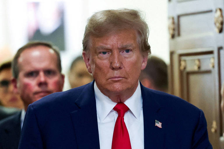 Foto mostra Donald Trump em primeiro plano. Ele é um homem idoso, branco, loiro, está de terno azul com gravata vermelha e tem um broche da bandeira americana na lapela. Sua expressão é de preocupação.